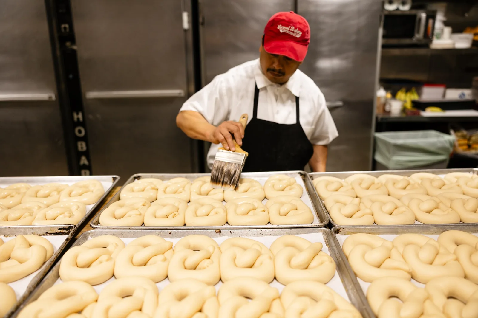A baker in a red cap working over trays of raw pretzels in the kitchen downstairs