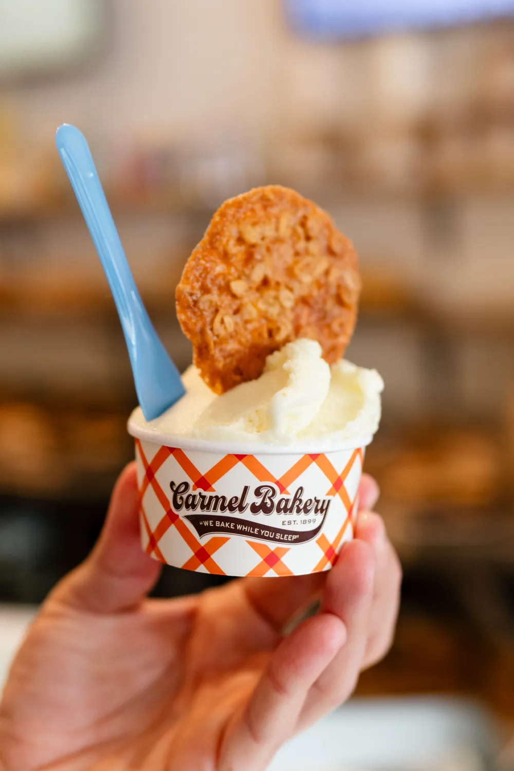 A hand holding a Carmel Bakery cup of gelato with a cookie in it, inside the shop