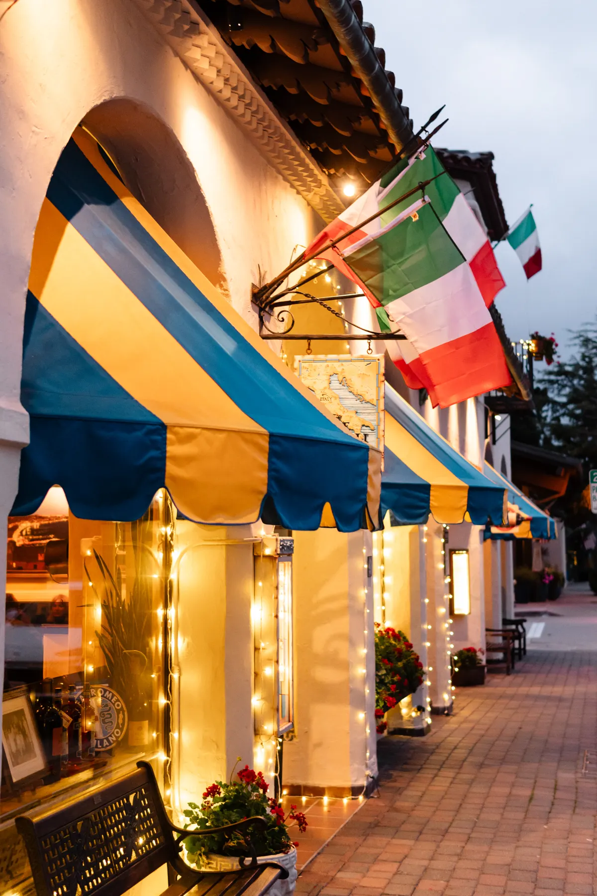 The Little Napoli storefront at dusk: blue and yellow awnings, Italian flags and string lights along Dolores Street