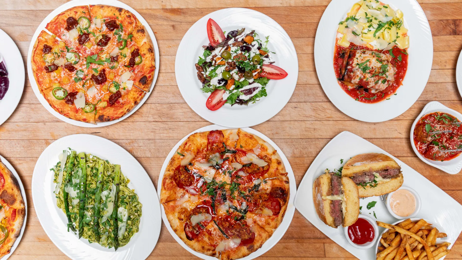 A table covered with Little Napoli plates seen from above: wood-fired pizzas, salads, parmigiana, asparagus and a sandwich with fries