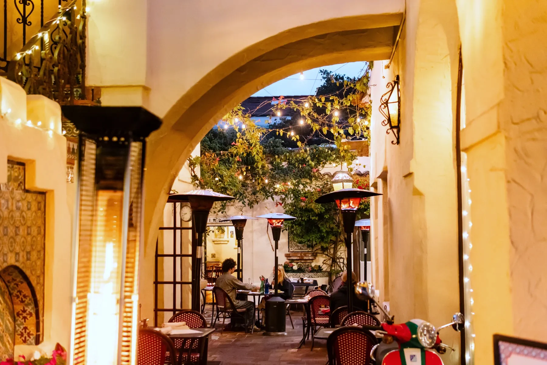 The Little Napoli courtyard at dusk through the El Paseo arch: guests at tables under heaters and string lights, a red Vespa by the wall