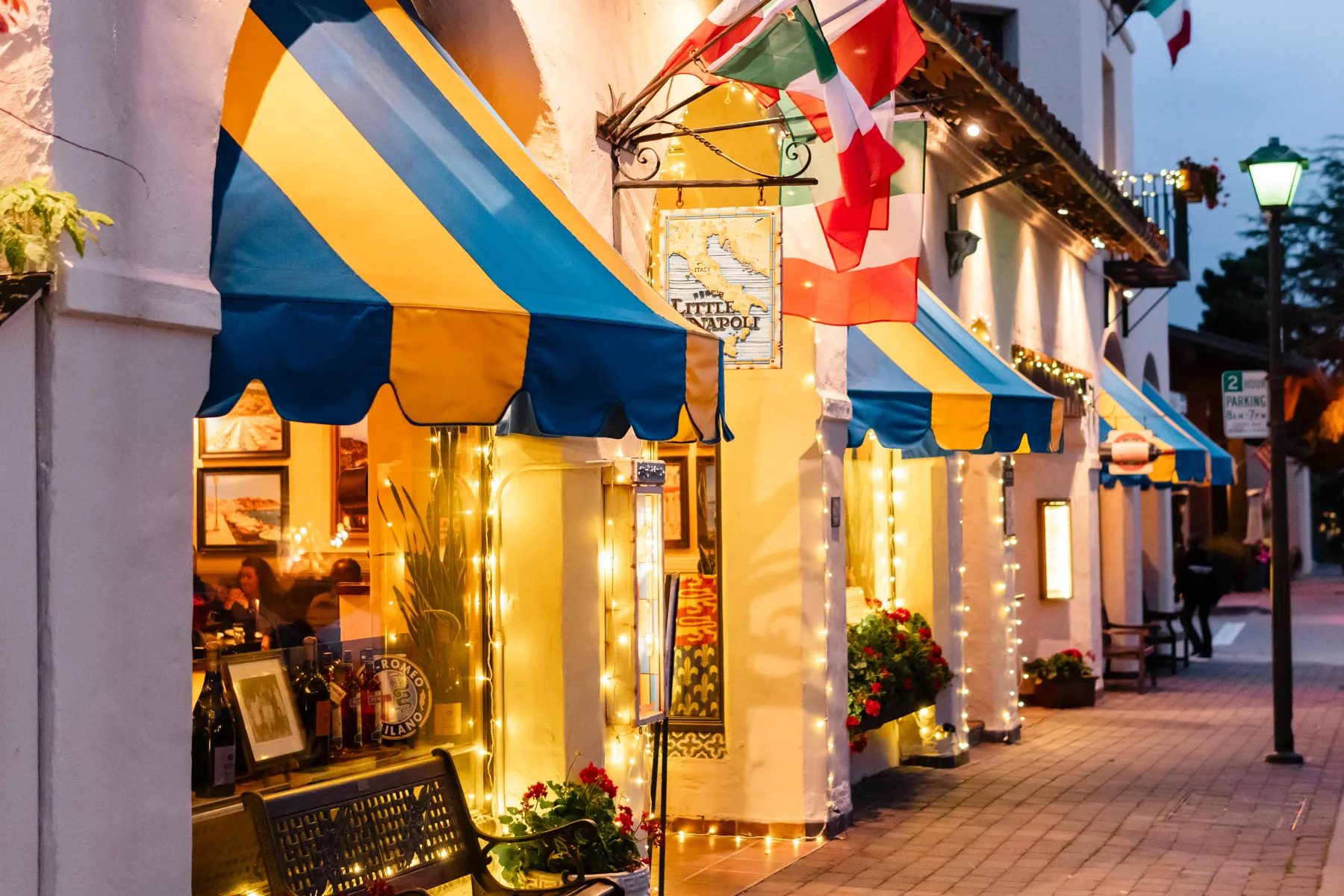 Blue and yellow striped awnings, Italian flags and string lights outside Little Napoli at dusk