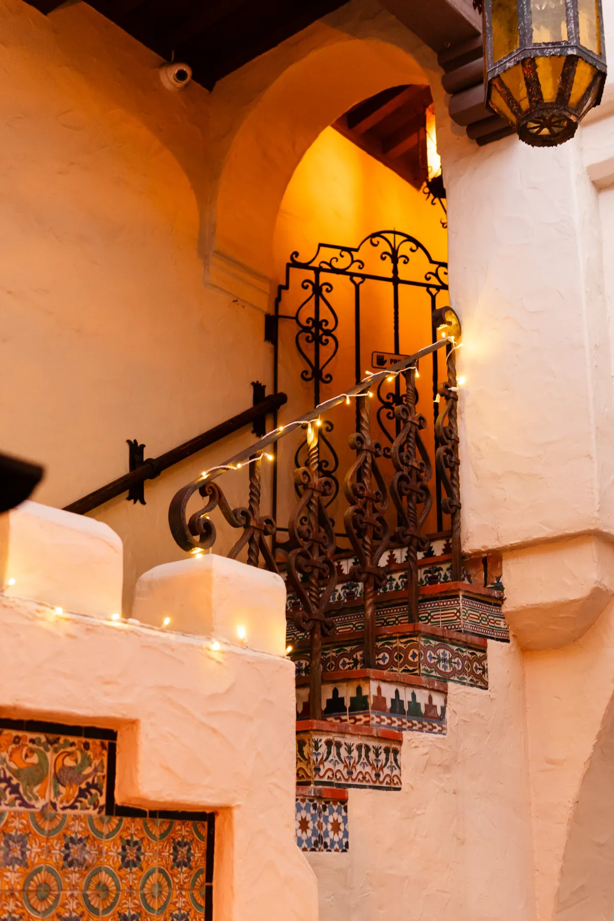 A white stucco stairway at the El Paseo Building with a wrought-iron gate and railing, patterned tile and string lights at golden hour